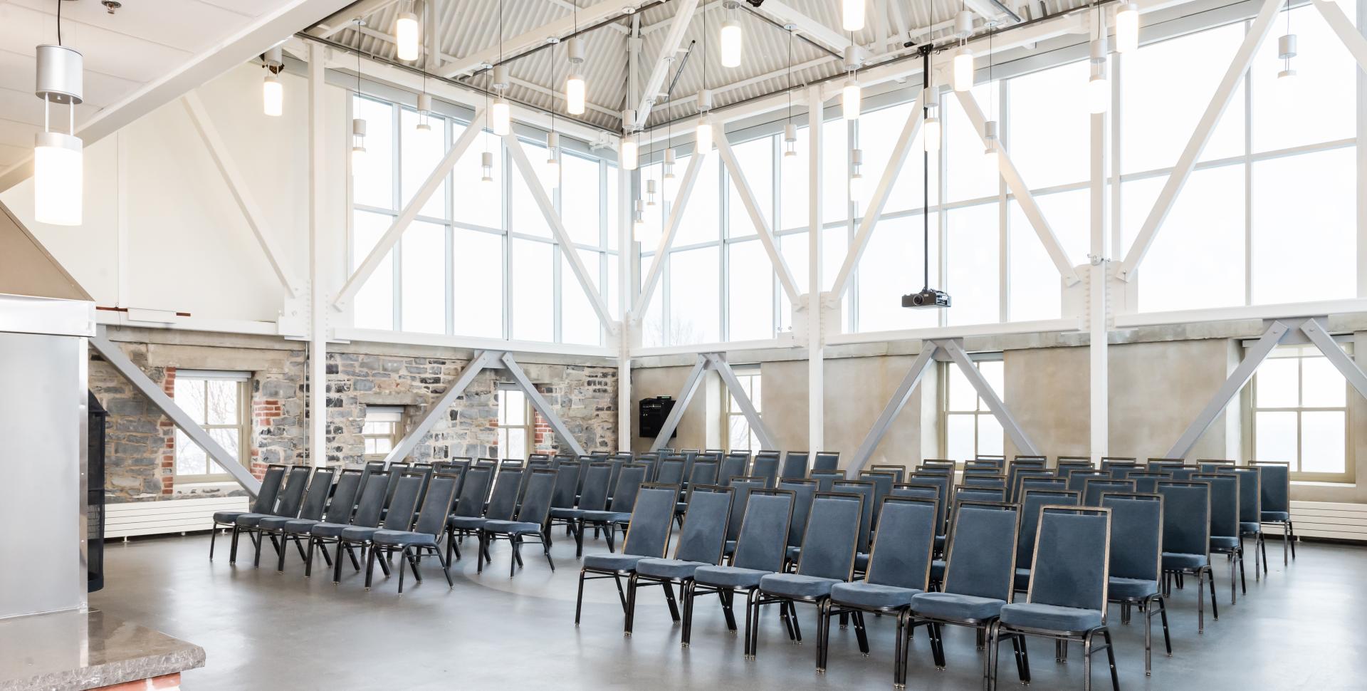 A large room with high ceilings and big windows, set with rows of chairs. .