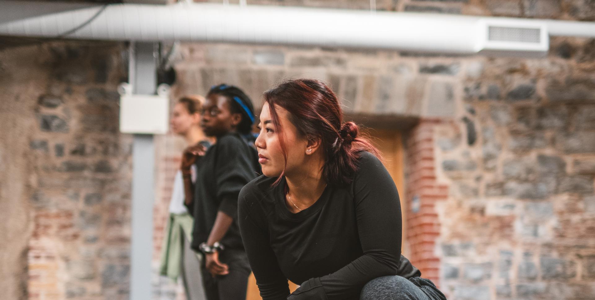 A dancer dressed in a black shirt and black pants kneels in a room with dark flooring and a stone wall. 