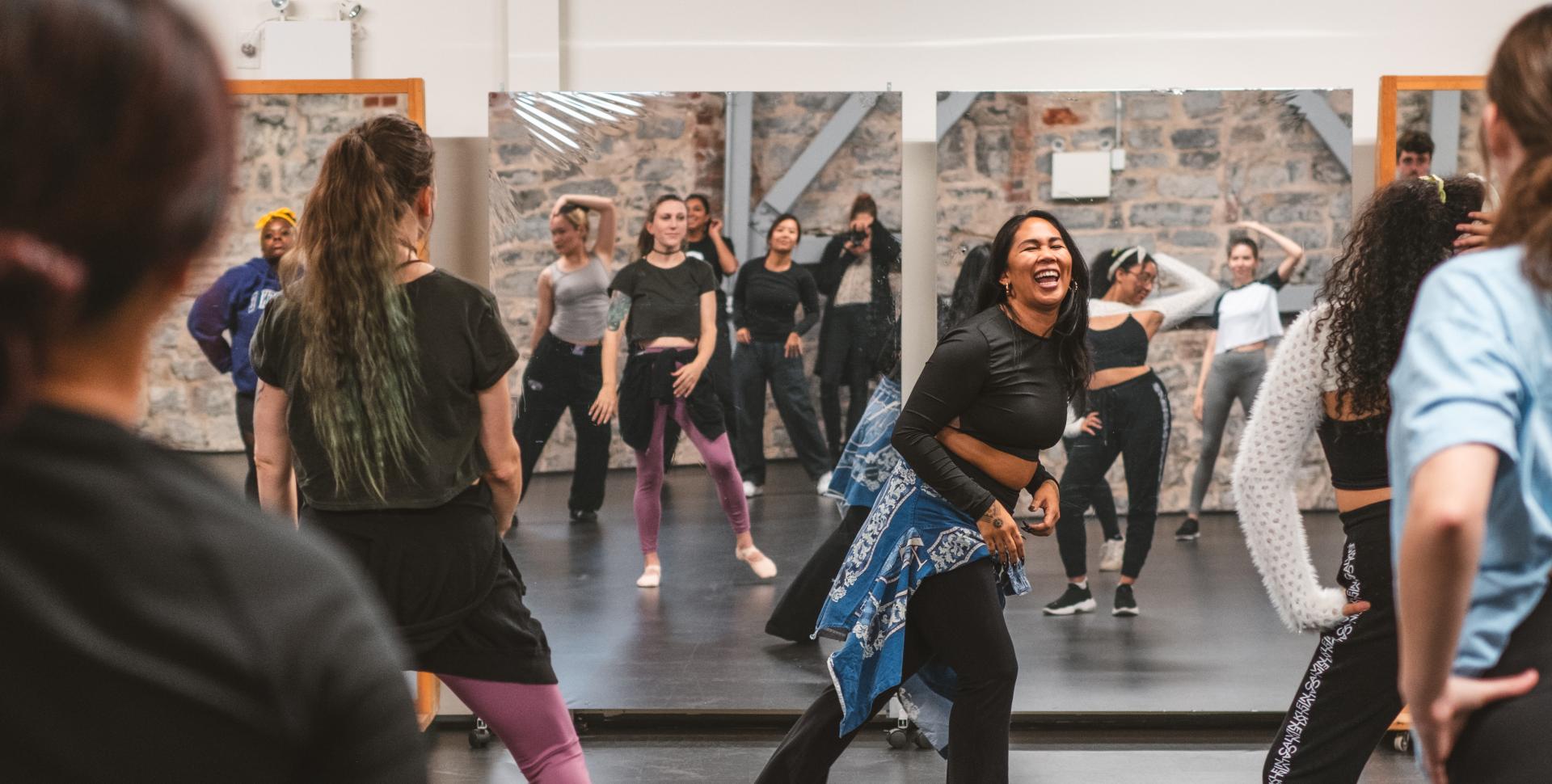 Dancers take a class in a room with black flooring and theatre lights overhead. 