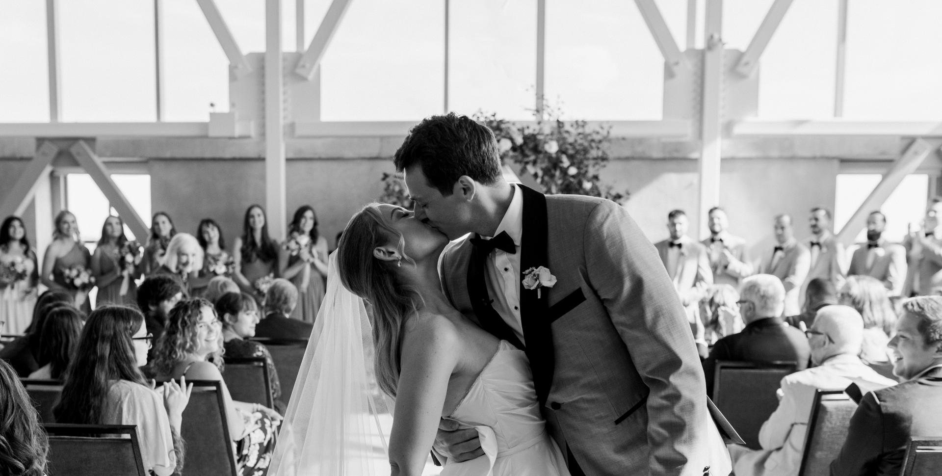 A newlywed couple kisses dramatically at the end of an aisle, in a room with large windows and white trusses. 