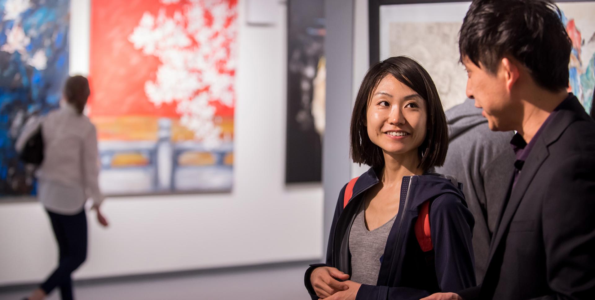 Two smiling people with dark hair and light brown skin smile and talk in front of a gallery space with large canvases. 