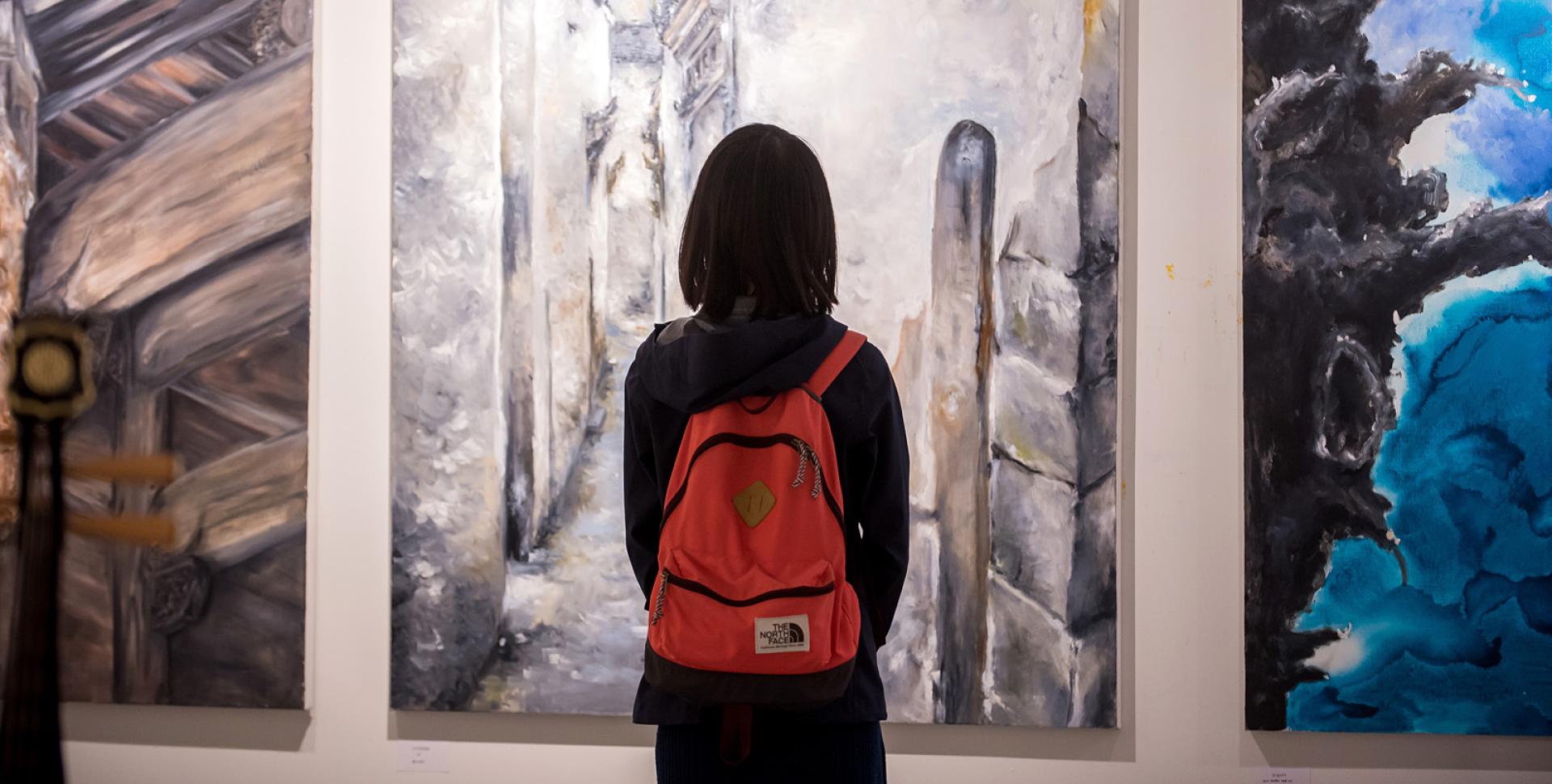 A person with dark hair and a red backpack takes in a large painting of an alleyway painted in warm tones. 