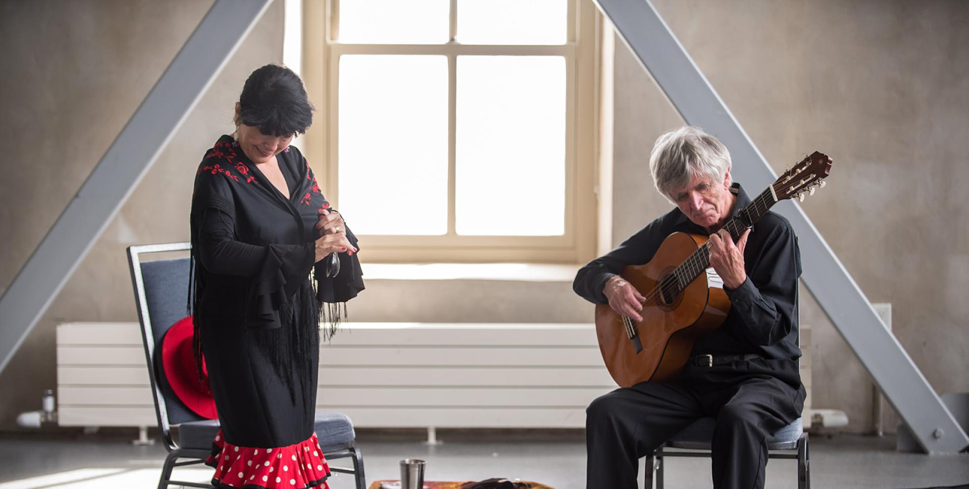 Two people in front of a large, bright window. On the right, a person with grey hair sits and plays the guitar. On the left, a person with dark hair and dark clothing with red accents dances. 