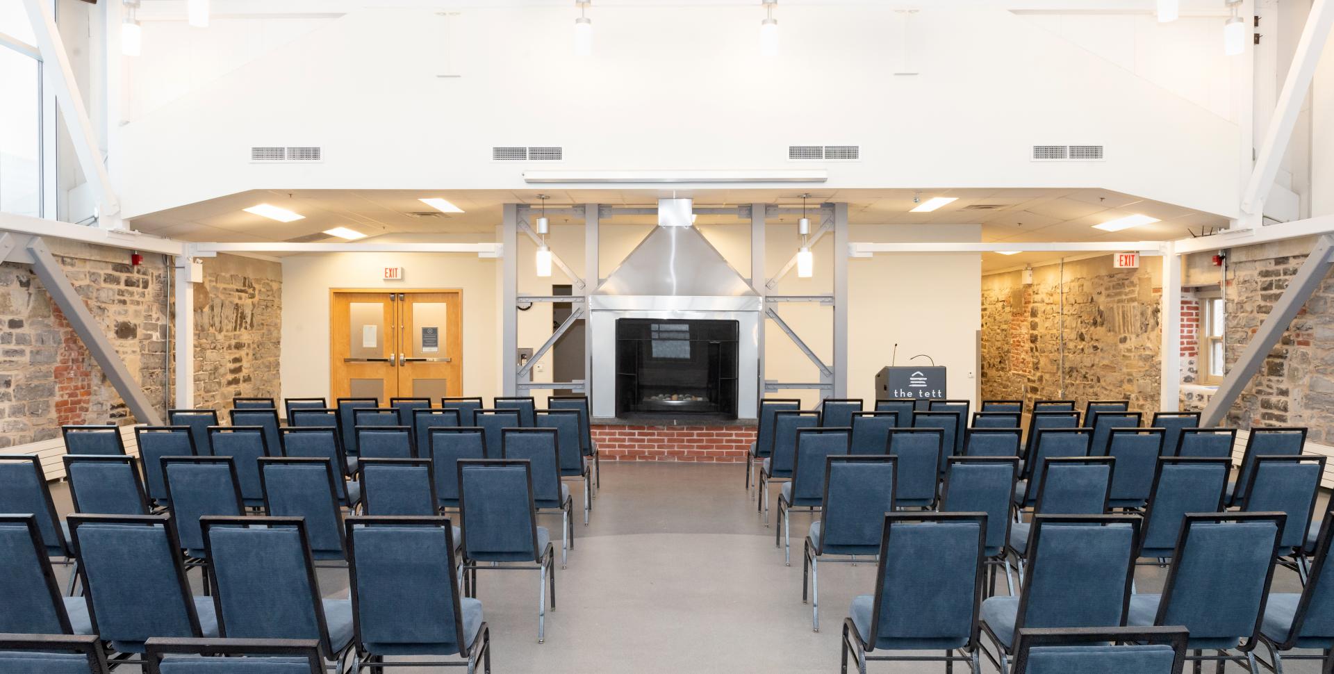 Rows of chairs in a large room face a large fireplace.