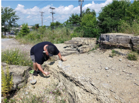 Sheldon bends over a large rock outdoors. 