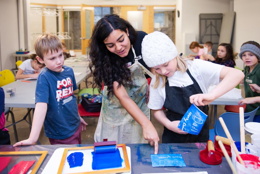 An instructor leads two kids on either side through a printmaking activity. 