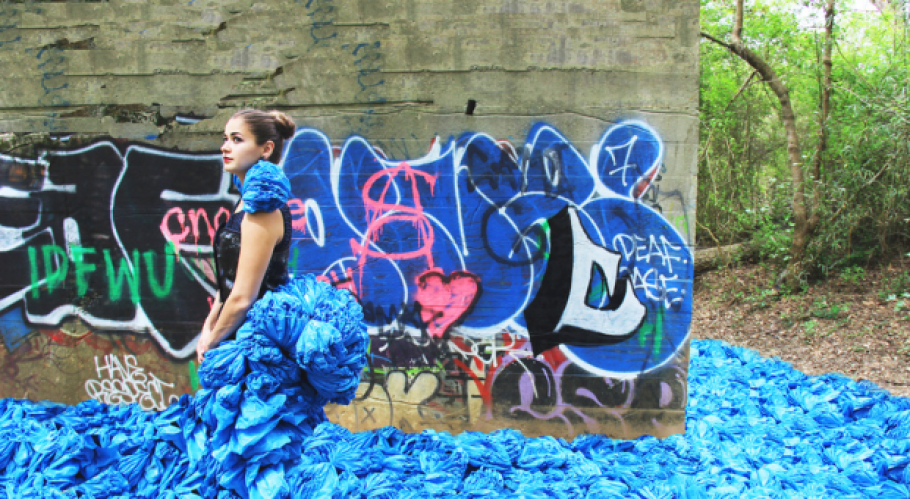  A person with brown hair in a bun wearing an haute couture outfit with blue fabric that cascades in front and behind like water, standing in front of graffiti. 