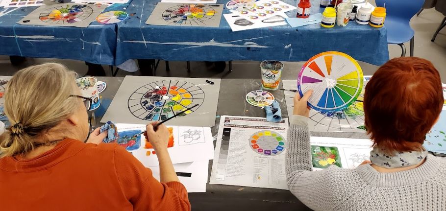 2 People sit in front of colourful paints, palettes, and colour wheels. 