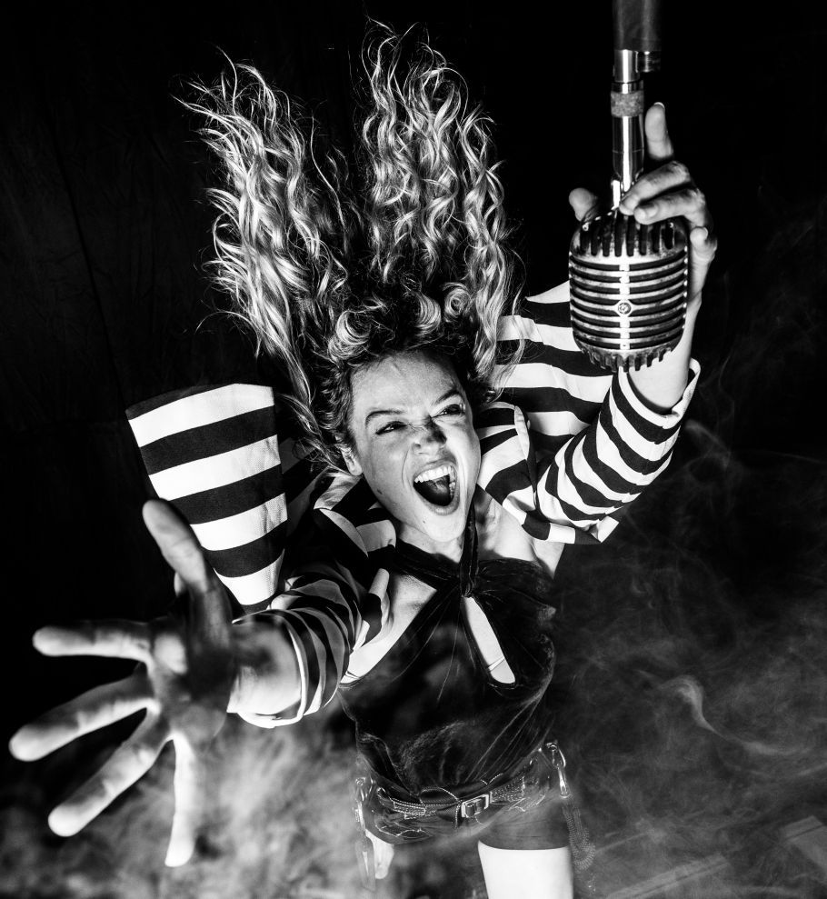 A woman wearing black and white, with long hair, jumps exuberantly towards a microphone. 