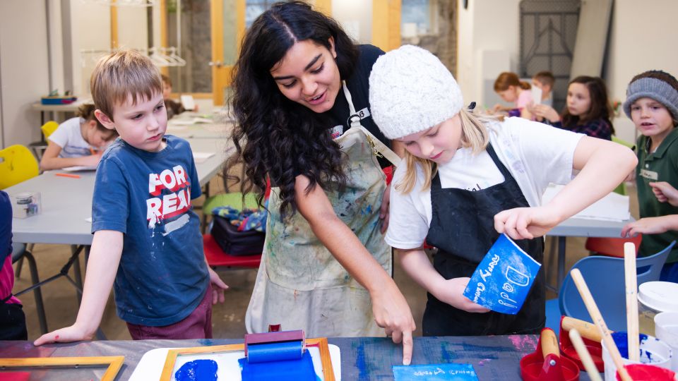 An instructor leads two kids on either side through a printmaking activity. 