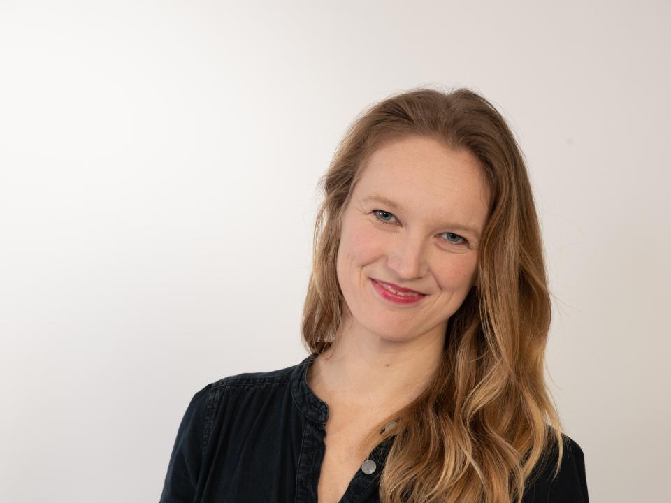 Jane Kirby, a white woman with long blonde hair and a jean dress, smiles at the camera. 