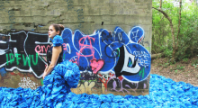  A person with brown hair in a bun wearing an haute couture outfit with blue fabric that cascades in front and behind like water, standing in front of graffiti. 